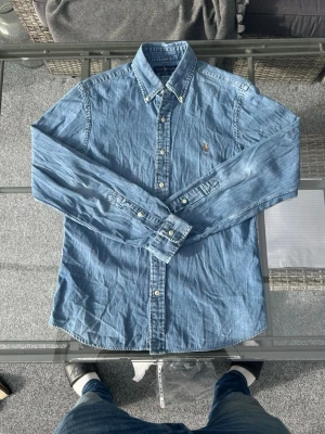 Ralph Lauren Denimskjorta – Slim Fit – M - Säljer en riktigt snygg denimskjorta från Ralph Lauren i modellen "Slim Fit". Skjortan har en klassisk tvättad blå nyans och den ikoniska hästloggan broderad på bröstet. Det här är ett perfekt basplagg som funkar till allt – bär den öppen över en t-shirt eller stängd för en mer dressad look. Skjortan är i storlek M och har en smickrande smal passform. Väldigt fint skick  Kort info:  Märke: Ralph Lauren  Modell: Slim Fit  Storlek: M  Färg: Blå (denim)  Skick: Mycket Fint