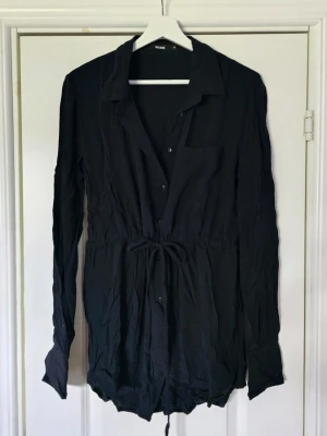 Svart skjortklänning med snörning - Svart skjortklänning från Bik Bok med långa ärmar, knappar framtill och snörning i midjan för en justerbar passform. Klassisk krage och bröstficka ger en stilren look. Perfekt till många olika stilar.