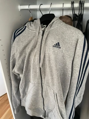 Grå zip hoodie från Adidas - Snygg grå hoodie från Adidas med dragkedja, blåa ränder längs ärmarna och Adidas-logga på bröstet. Tillverkad i mjuk bomullsblandning (70% bomull, 30% polyester). Praktisk huva med dragsko och två fickor framtill.
