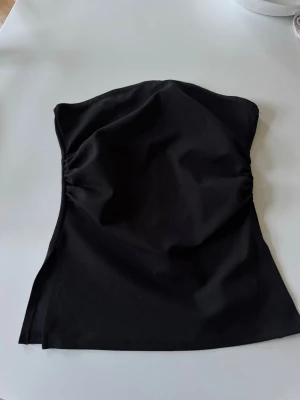 Svart axelbandslös tubtopp - Snygg svart axelbandslös tubtopp med enkel och stilren design. Toppen har en lätt figurnära passform och är perfekt att matcha med både jeans och kjol. Materialet känns mjukt och följsamt.