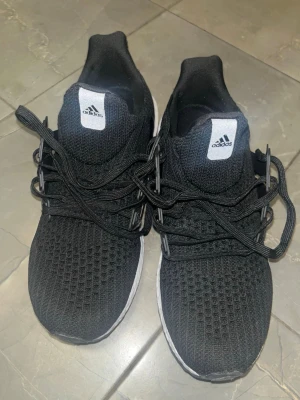 Svarta Adidas Ultra Boost sneakers - Svarta Adidas Ultra Boost sneakers med vit sula och klassisk BOOST-dämpning. Ovandelen är i ventilerande mesh och skorna har svarta snören. Perfekta för dig som gillar sportig och stilren look.