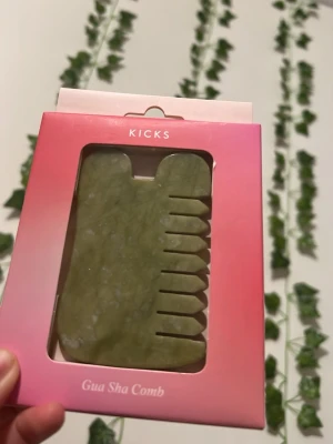 Grön Gua Sha kam från Kicks - En grön Gua Sha kam från Kicks, tillverkad i slät sten. Kammen har en unik form med rundade kanter och tänder på ena sidan, perfekt för ansiktsmassage och hårbottenmassage. Kommer i en rosa förpackning.