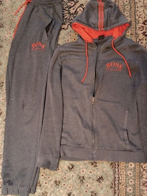 Grå loungewear-set från Hugo Boss - Mysigt loungewear-set från Hugo Boss i grått med röda detaljer. Hoodien har dragkedja, röda snören och röd insida på huvan samt BOSS-logga på bröstet. Matchande byxor med resår i midjan och BOSS-tryck. Tillverkat i mjuk bomullsblandning för maximal komfort. Använd 4/5 gånger den hsr fdn bars legat där. Inga Hål eller skador på både byxorna och koftan