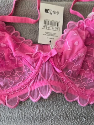 Rosa spets-bh med hjärtmönster - Supersöt rosa bh från Body med transparent spets och broderade hjärtan. Dekorerad med en liten rosett framtill och vågig kant. Justerbara axelband och hakknäppning bak. Helt ny med prislapp