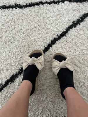 Beiga sandaler med rosett - Säljer ett par beiga sandaler med stor tygrosett framtill och flätad platåsula i jute. Sandalerna har öppen tå och en trendig, avslappnad look som passar perfekt till sommaren. Materialet är tyg och sula i repstil för en bohemisk vibe.