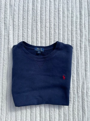 Mörkblå sweatshirt från Polo Ralph Lauren - Klassisk mörkblå sweatshirt från Polo Ralph Lauren med rund halsringning och röd broderad logga på bröstet. Tröjan har långa ärmar och ribbade muddar vid ärmslut och nederkant. Perfekt för en avslappnad och stilren look.