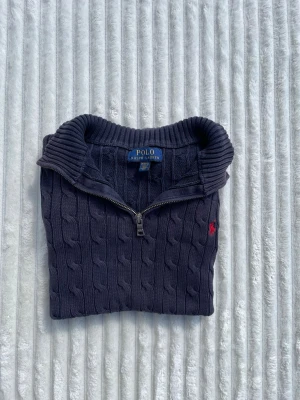 Mörkblå kabelstickad tröja Polo Ralph Lauren - Mörkblå kabelstickad tröja från Polo Ralph Lauren med halv dragkedja och ribbad krage. Tröjan har ett rött broderat logomärke på bröstet och ribbade muddar vid ärmslut och nederkant. Perfekt för en stilren och klassisk look.