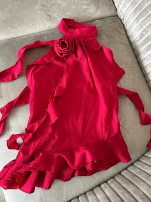 Röd omlottopp med volang och rosett - Snygg röd omlottopp med hög krage, dekorativ blomrosett vid halsen och volangkant nedtill. Toppen har knytband i sidorna och är ärmlös, vilket ger en chic och trendig look. Perfekt för dig som vill sticka ut med en färgstark och unik topp.
