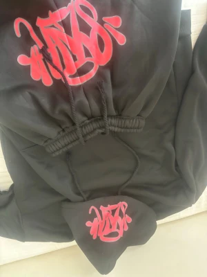 Syna World - Säljer ett svart loungewear-set med hoodie och byxor. Hoodien har en stor ficka framtill och coolt rött graffiti-tryck på både bröstet och huvan. Byxorna har resår i midjan och dragsko för skön passform. Perfekt för chill dagar hemma.