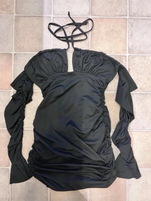 Svart kort klänning med snörning - Trendig svart kort klänning med långa ärmar och snygg snörning i halsen. Klänningen har rynkade detaljer längs sidorna och en unik offshoulder-design. Perfekt för dig som vill sticka ut med en cool och modern look.