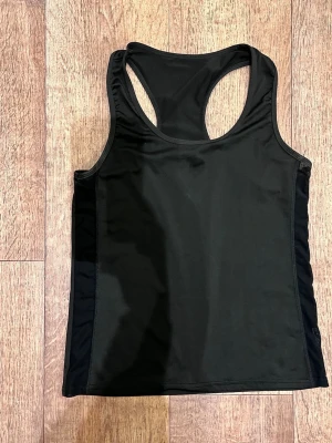 Svart träningstopp i syntet - Snygg svart träningstopp med brottarrygg, perfekt för gymmet eller löprundan. Tillverkad i ett stretchigt syntetmaterial som andas och sitter skönt mot kroppen. Enkel och stilren design utan mönster eller logga.