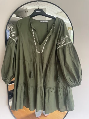 Grön boho-klänning från Zara - Säljer en grön boho-inspirerad kortklänning från Zara med broderade vita detaljer och volanger. Klänningen har v-ringning med snörning, vida långärmar och avslappnad passform. Perfekt för sommarens alla tillfällen!