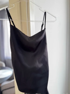 Svart satin topp med knytning i ryggen - Stilren svart topp i satin med tunna justerbara axelband och snygg knytning i ryggen. perfekt för dig som gillar minimalistisk och trendig stil.