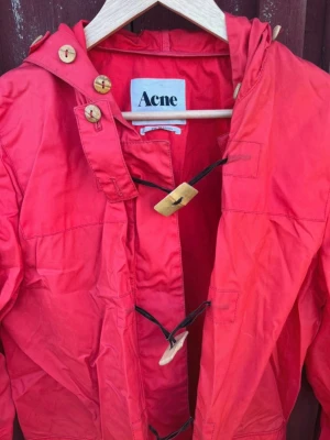 Vintage Röd regnjacka från Acne med träknappar - Snygg röd regnjacka från Acne med huva och träknappar som stängning. Jackan har stora fickor framtill och detaljerade knappar i trä och plast. Lång modell med avslappnad passform, perfekt för regniga dagar. Materialet är tunt och vattenavvisande.