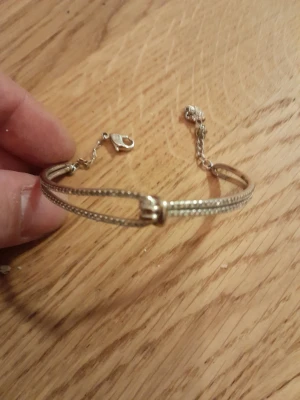 Silvrigt armband med knutdetalj - Snyggt Swarowski armband i silverfärg med en unik knutdetalj framtill. Armbandet har en tunn kedja med justerbar längd och stängs med ett klassiskt klolås. Perfekt accessoar för att lyfta din stil med en enkel men elegant touch.