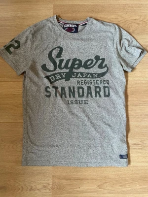 Grå Superdry t-shirt med tryck - Snygg grå t-shirt från Superdry med stort tryck på bröstet och siffra på ärmen. Klassisk passform och rund halsringning. Perfekt för en avslappnad stil och enkel att matcha med jeans eller shorts.
