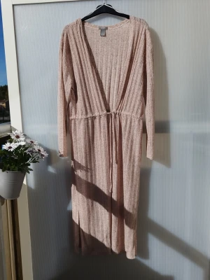 Beige ribbad lång kofta - Beige lång kofta i ribbat material med djup V-ringning och knytband i midjan. Koftan har långa ärmar och ett mjukt, stretchigt tyg som ger en avslappnad och trendig look.