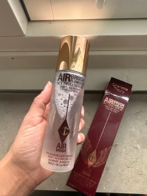 Charlotte Tilbury Airbrush Setting Spray - Airbrush Flawless Setting Spray från Charlotte Tilbury i en genomskinlig flaska med guldfärgat lock. Ger en fräsch finish och håller sminket på plats hela dagen. Flaskan har elegant design med vingar och logga i guld. Perfekt för att fixera makeup och få en naturlig look.