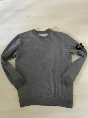 Grå sweatshirt från Stone Island - Snygg grå sweatshirt från Stone Island med klassisk rund hals och ribbade muddar. Tröjan har den ikoniska Stone Island-loggan på vänster ärm. Tillverkad i mjukt bomullsmaterial som är skönt mot huden. Perfekt för en avslappnad och stilren look. Storlek S/M