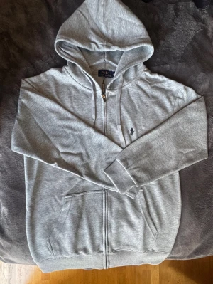Grå zip hoodie från Polo Ralph Lauren - Snygg grå zip hoodie från Polo Ralph Lauren med klassisk logga broderad på bröstet. Hoodien har dragkedja, två fickor framtill och justerbar huva med snören. Tillverkad i mjukt bomullsmaterial för skön komfort och avslappnad stil.