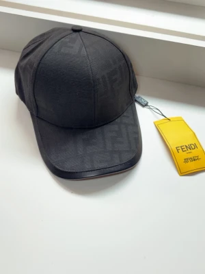 Svart keps från Fendi med logga - Snygg svart keps från Fendi med diskret FF-monogram över hela tyget. Klassisk böjd skärm med läderdetalj längs kanten och justerbar passform bak. Kepsen har en stilren och modern look, perfekt för dig som gillar exklusiva accessoarer.
