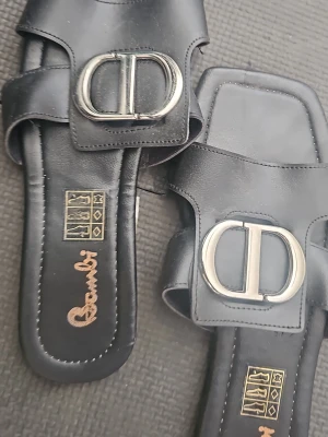Svarta sandaler från Bambi med metallspänne - Stilrena svarta sandaler från Bambi med bred rem och stort silverfärgat metallspänne framtill. Sandalerna har fyrkantig tå och är tillverkade i slätt konstläder. Perfekta för en trendig och avslappnad look.