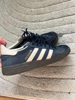 Adidas Spezial mörkblå sneakers - Säljer ett par Adidas Spezial sneakers i mörkblå mocka med vita detaljer och klassiska tre ränder på sidan. Skorna har svart sula, vita hälpartier och guldfärgad Spezial-text. Perfekta för dig som gillar retrostil och streetwear.
