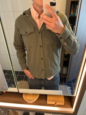 Olivgrön overshirt från Jack & Jones - Olivgrön overshirt från Jack & Jones med två bröstfickor med lock och knappar framtill. Tillverkad i mjukt tyg som ger en avslappnad look. Perfekt att bära över en skjorta eller t-shirt för en snygg lager-på-lager stil.