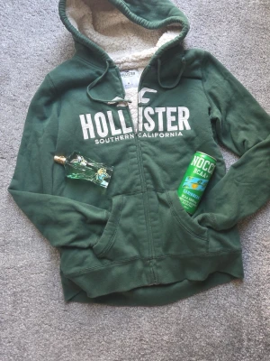 Hollister hoodie - Mysig grön hoodie från Hollister med vit logga och texten 'Southern California' på bröstet. Utrustad med dragkedja, två fickor och en fluffig, vit foder i huvan. Pris kan diskuteras.💯🫱🏻‍🫲🏽