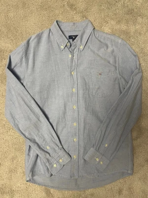 Ljusblå skjorta från GANT - Klassisk ljusblå skjorta från GANT med button-down krage och bröstficka med liten logga. Skjortan har långa ärmar och stängs med ljusa knappar framtill. Perfekt till jeans eller chinos för en clean look.