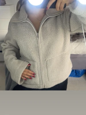 Beige teddyjacka med dragkedja - Mysig beige teddyjacka med dragkedja framtill och hög krage. Jackan har två stora fickor och är tillverkad i fluffigt, mjukt teddy-material.