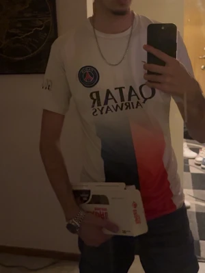 PSG matchtröja vit med tryck - Säljer en vit PSG matchtröja med Qatar Airways-tryck på bröstet och klubbmärke på vänster sida. Tröjan har en snygg design med blå och röda fält framtill och korta ärmar. Perfekt för dig som älskar fotboll och vill stötta ditt favoritlag.