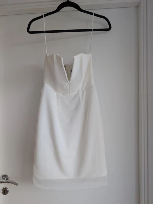 Vit kort klänning bandeau - Stilren vit kort bandeauklänning från Rebecca Stella med djup cut out v-ringning framtill. Klänningen har en figurnära passform och dragkedja i ryggen. Perfekt för sommarens alla tillfällen!