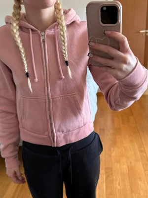 Rosa hoodie med dragkedja och huva - Snygg rosa hoodie med dragkedja framtill, två fickor och justerbar huva med dragsko. Tillverkad i mjukt sweatshirtmaterial och har ribbade muddar vid ärmslut och nederkant. Enkel att styla till olika looks.