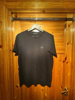 Svart t-shirt från Acne Studios - En stilren svart t-shirt från Acne Studios med klassisk passform och rund hals. T-shirten har korta ärmar och en liten diskret logopatch på bröstet. Perfekt basplagg för alla tillfällen. Köpare står för frakt 📦 