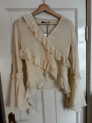 Beige volangblus från BikBok - Supersöt beige blus från BikBok med omlott-look, volangdetaljer och v-ringning. Blusen har långa, vida ärmar med volanger och är gjord i ett lätt, skirt tyg som ger en romantisk vibe. Perfekt att styla med jeans eller kjol för en trendig look. Aldrig använd och har prislappen kvar. 💕