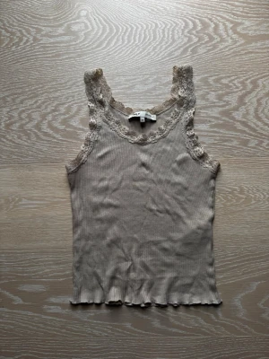 Beige ribbad linnetopp med spets - Säljer en beige ribbad linnetopp från Only med fina spetsdetaljer runt halsringningen och axelbanden. Toppen är ärmlös och har en lätt vågig kant nedtill. Perfekt att styla med jeans eller kjol för en enkel men söt look.