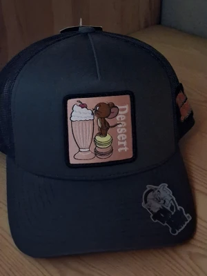 Tom & Jerry Keps  - Cool svart keps med nät på baksidan och patch framtill med Jerry, milkshake och macarons samt texten 'Dessert'. Extra Tom & Jerry-märke på skärmen och sidan. Justerbar storlek och böjd skärm. Perfekt för dig som gillar tecknade klassiker.
