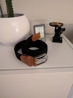 Svart flätat bälte med bruna detaljer - Snyggt svart flätat bälte med bruna skinn-detaljer och silverfärgat spänne. Perfekt för att lyfta vilken outfit som helst. Längd 95 cm. Passar både jeans och chinos.