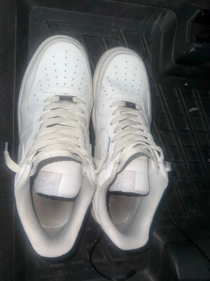 Nike Air Force 1 vita sneakers - Klassiska Nike Air Force 1 sneakers i helvitt läder med perforerad tåbox och vita skosnören. Ikonisk design med Nike-logga på plösen och sidan. Skorna har en chunky sula och tidlös streetstil som alltid funkar.