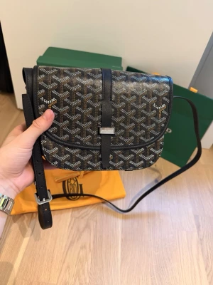 Svart axelväska från Goyard - Snygg svart axelväska från Goyard med det klassiska vita och gråa mönstret. Väskan har justerbar axelrem, silverfärgad spänndetalj och rundad form. Tillverkad i canvas med läderdetaljer. Perfekt för dig som vill ha något trendigt och exklusivt.