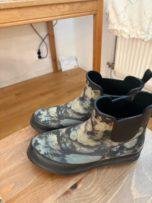 Blommiga Chelsea boots i gummi - Säljer ett par Chelsea boots i gummi med blommigt mönster i blått, grönt och vitt. Skorna har svarta elastiska paneler på sidorna och dragflikar både fram och bak. Perfekta för regniga dagar och ger en cool touch till din outfit.