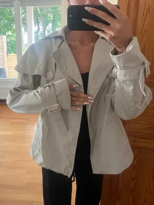 Ljusbeige kort trenchcoat - Kort trenchcoat i ljusbeige med bred krage, dubbelknäppning och dekorativa knappar. Jackan har bälte vid ärmsluten och en avslappnad passform. 