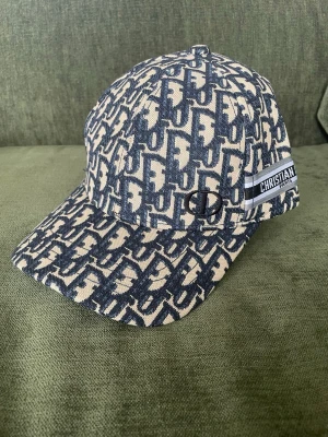 Beige och blå keps från Dior - Säljer en snygg keps från Christian Dior med beige och blått monogrammönster. Kepsen har böjd skärm, broderad logga på sidan och ett band med texten 'Christian Dior Paris'. Tillverkad i ett slitstarkt tyg med exklusiv känsla.