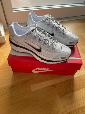 Nike P6000  - Nike P-6000 sneakers i vitt med svarta detaljer och silvriga inslag. Skorna har en sportig design med mesh och syntetmaterial, samt den klassiska svarta swoosh-loggan på sidan. Perfekta för dig som gillar retrostil och streetwear.