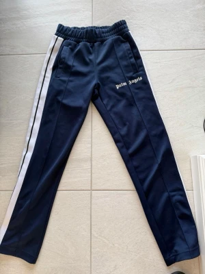 Palm Angels blå mjukisbyxor - Palm Angels mjukisbyxor i mörkblått med vita sidoränder och logga på vänster ben. Byxorna har resår i midjan och raka ben. Tillverkade i ett mjukt och stretchigt material som känns skönt mot huden. Perfekta för en avslappnad och trendig look.