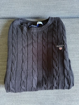 Svart kabelstickad tröja GANT - Kabelstickad tröja från GANT i mörkblått med rund halsringning och broderad logga på bröstet. Klassisk och stilren design med ribbade muddar. Perfekt för dig som gillar preppy stil.