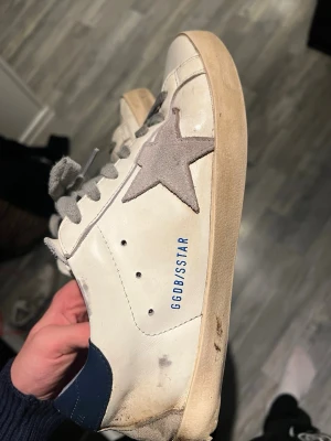 Golden Goose vita sneakers med grå stjärna - Säljer ett par vita Golden Goose sneakers med grå mockastjärna på sidan och blå detaljer vid hälen. Skorna har gråa snören och är tillverkade i läder med inslag av mocka. Klassisk låg modell med vintage-inspirerad look och GGDB/SSTAR-tryck på sidan.