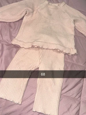 Ljusrosa ribbat set med volangkant - Säljer ett ljusrosa ribbat set med långärmad tröja och matchande byxor. Tröjan har en söt volangkant nedtill och båda delarna är i mjukt, stretchigt material. Perfekt för en mysig och stilren look.