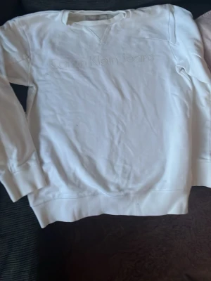 Vit sweatshirt från Calvin Klein Jeans - Säljer en stilren vit sweatshirt från Calvin Klein Jeans med diskret broderad logga på bröstet. Tröjan har rund halsringning, långa ärmar och ribbade muddar vid ärmslut och nederkant. Perfekt för en clean och avslappnad look.